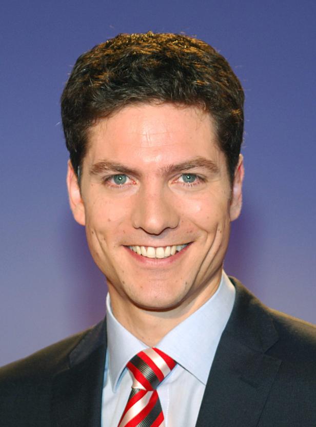 Ein lächelnder Mann mit Anzug und Krawatte vor blauem Hintergrund.