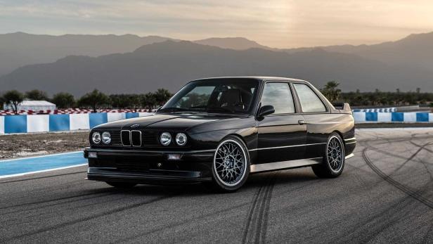 Ein schwarzer BMW E30 steht auf einer Rennstrecke vor einer Bergkulisse.