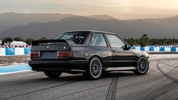 Ein schwarzer BMW E30 steht auf einer Rennstrecke vor einer Bergkulisse.
