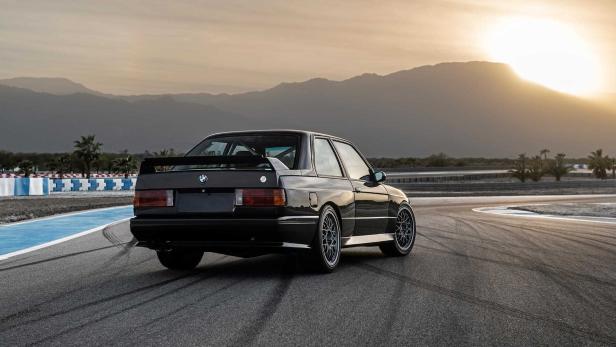 Ein schwarzer BMW E30 steht auf einer Rennstrecke vor einer Bergkulisse.