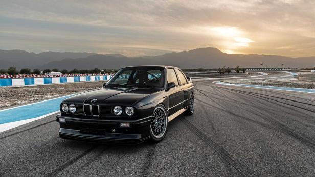 Ein schwarzer BMW E30 steht auf einer Rennstrecke vor einer Bergkulisse.