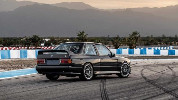 Ein schwarzer BMW E30 steht auf einer Rennstrecke vor einer Bergkulisse.