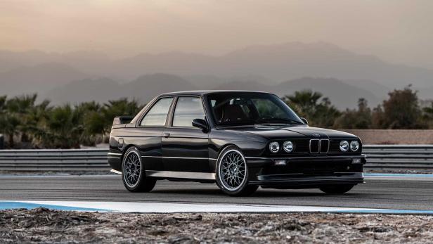 Ein schwarzer BMW E30 steht auf einer Rennstrecke vor einer Bergkulisse.