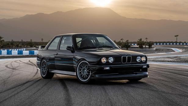 Ein schwarzer BMW E30 steht auf einer Rennstrecke vor einer Bergkulisse.