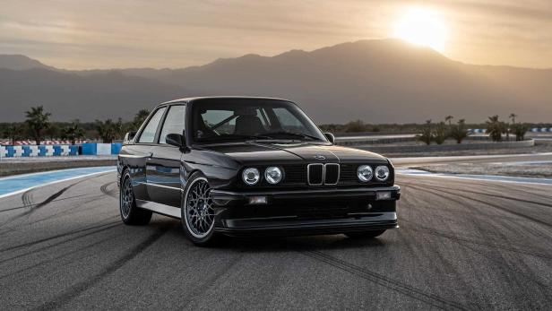 Ein schwarzer BMW E30 steht auf einer Rennstrecke vor einer Bergkulisse.