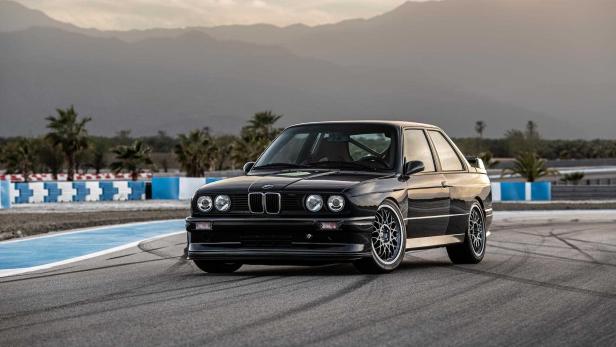 Ein schwarzer BMW E30 steht auf einer Rennstrecke vor einer Bergkulisse.