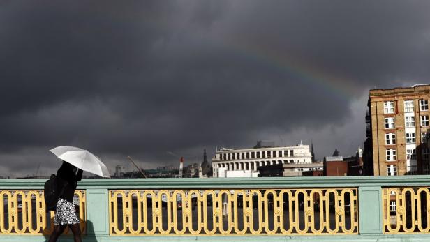 Eine Person mit Regenschirm geht bei dunklem Himmel und Regenbogen über eine Brücke.
