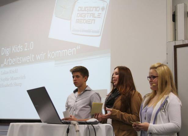 Drei junge Leute präsentieren „Digi Kids 2.0“ vor einem Publikum.