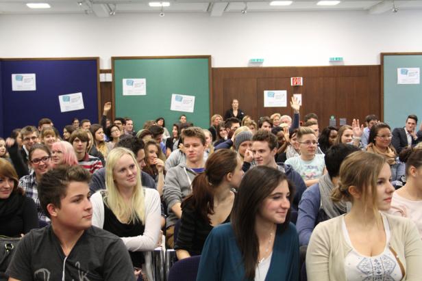 Ein Publikum sitzt in einem Raum, einige Leute heben die Hände.