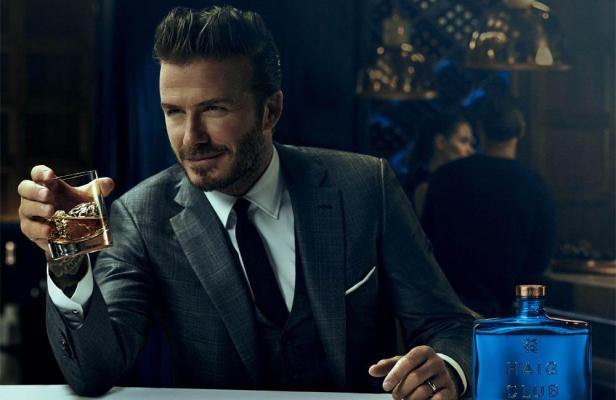 David Beckham in einem grauen Anzug hält ein Glas Whisky in einer Bar.