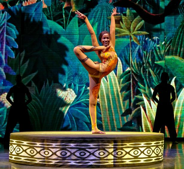 Eine Akrobatin in einem orangefarbenen Kostüm steht auf einem Bein auf einer Bühne vor einem Dschungelhintergrund.