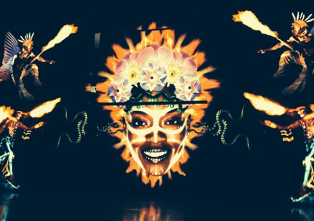 Eine Frau mit Blumenkrone und Gesichtsbemalung, umgeben von Figuren mit Flügeln und Fackeln.