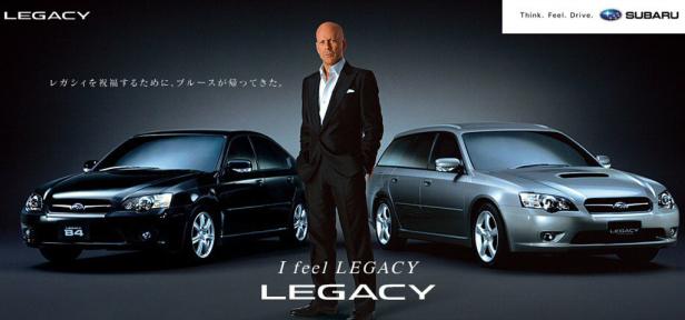 Bruce Willis posiert zwischen einem schwarzen Subaru Legacy B4 und einem silbernen Subaru Legacy.