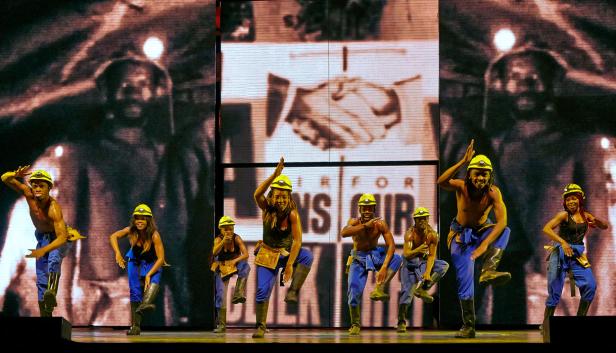 Eine Gruppe von Tänzern in Arbeitskleidung und Helmen tritt auf einer Bühne vor einer Projektion auf.