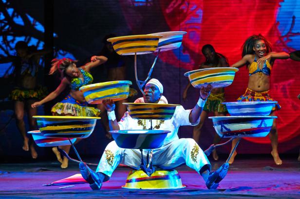 Ein Mann balanciert mehrere bemalte Schüsseln auf seinem Körper, während Tänzerinnen im Hintergrund auftreten.