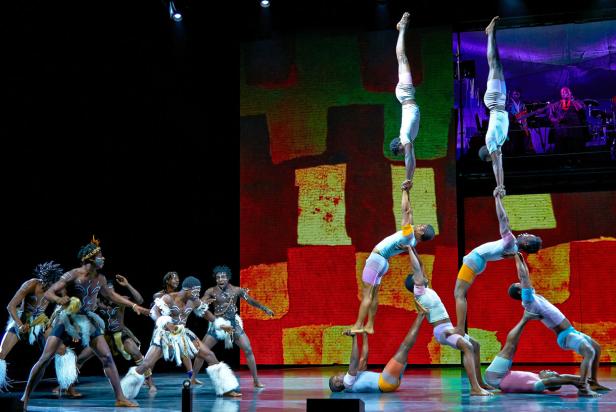 Eine Gruppe von Akrobaten und Tänzern tritt auf einer Bühne vor einem farbenfrohen Hintergrund auf.