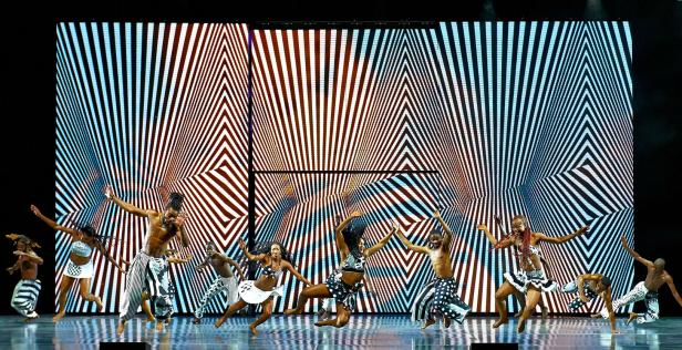 Eine Gruppe von Tänzern tritt vor einem abstrakten, geometrischen Hintergrund auf.