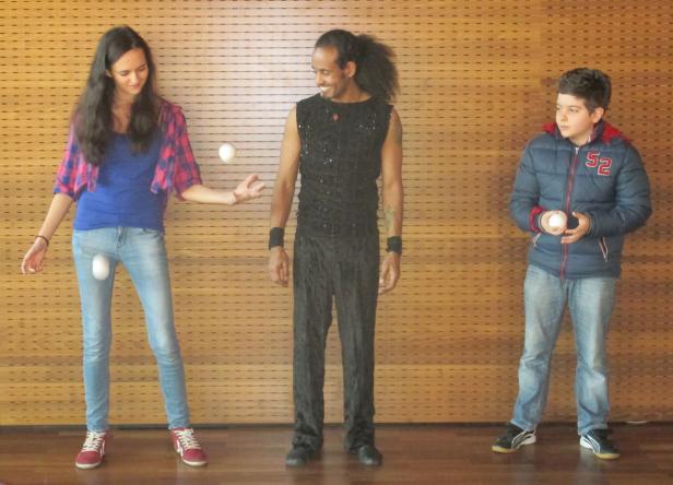 Eine Frau, ein Mann und ein Junge jonglieren mit weißen Bällen vor einer Holzwand.