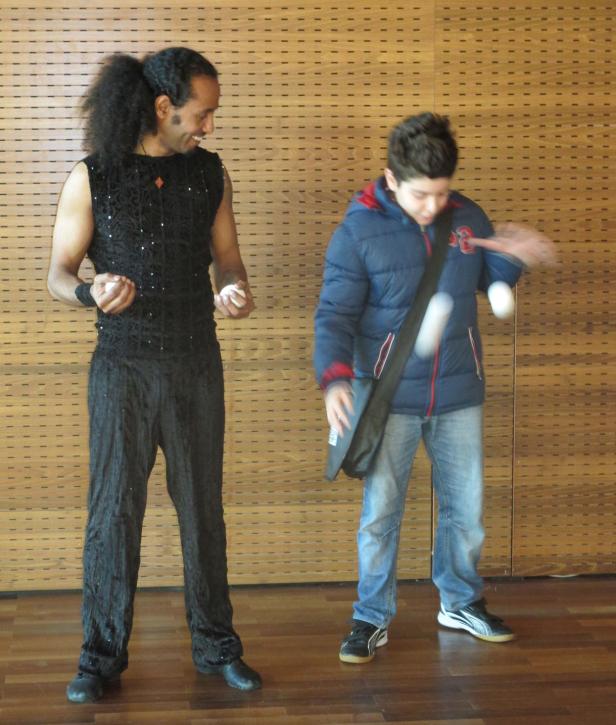 Ein Mann im schwarzen Outfit und ein Junge jonglieren mit weißen Bällen.