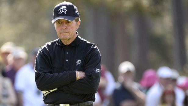 Ein Mann mit Kappe und verschränkten Armen steht auf einem Golfplatz.