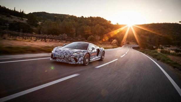 Ein mit Mustern beklebter McLaren fährt auf einer kurvenreichen Straße in Richtung Sonnenuntergang.
