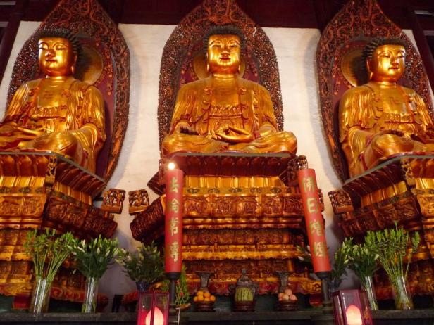 Drei goldene Buddha-Statuen sitzen auf verzierten Podesten in einem Tempel.