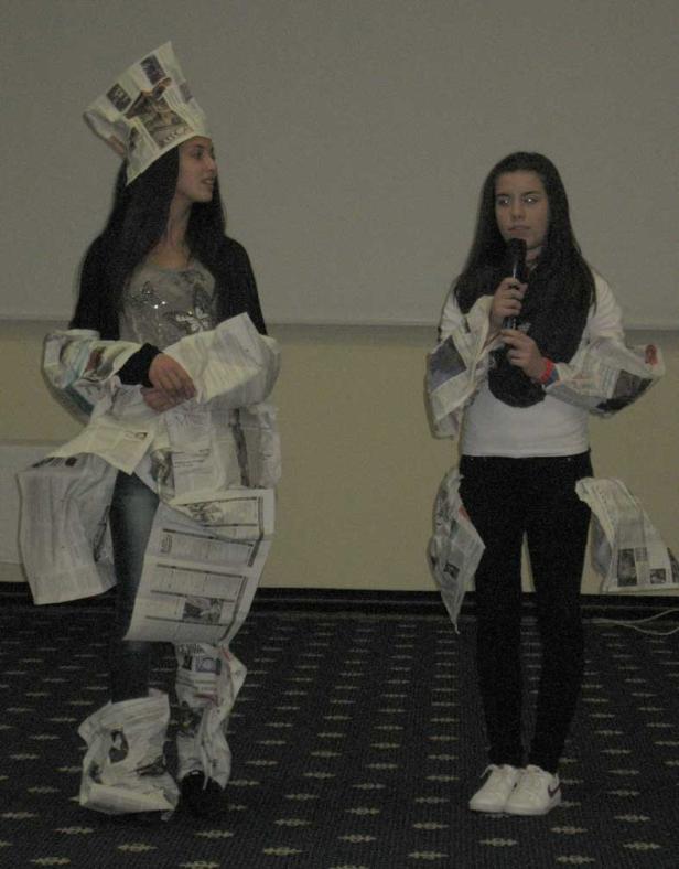 Zwei junge Frauen präsentieren Kleidung aus Zeitungspapier, eine spricht ins Mikrofon.