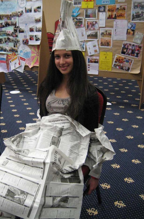 Eine junge Frau sitzt mit einem Kleid und einem Hut aus Zeitungspapier da.