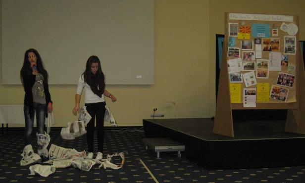 Zwei junge Frauen stehen auf einer Bühne, eine spricht ins Mikrofon, die andere wirft Zeitungspapier in die Luft.