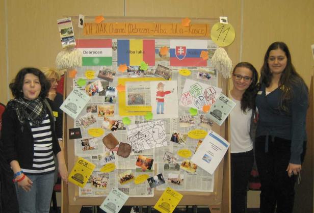 Drei junge Frauen stehen vor einer Pinnwand mit Fotos und Flaggen von Debrecen, Alba Iulia und Košice.