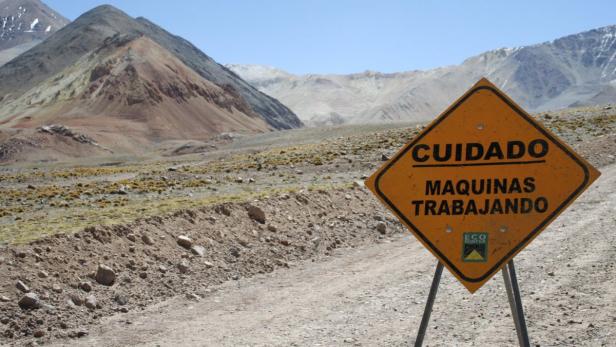 Ein gelbes Warnschild mit der Aufschrift „Cuidado Maquinas Trabajando“ steht vor einer Berglandschaft.