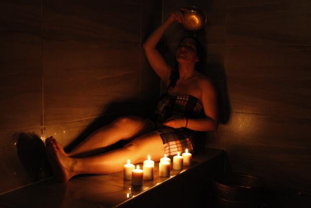 Eine Frau gießt Wasser aus einer Schale über sich in einem von Kerzen beleuchteten Raum.