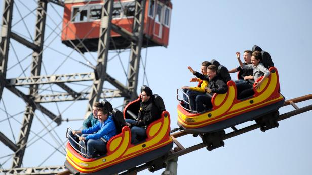 Zwei Wagen einer Achterbahn fahren vor dem Wiener Riesenrad.
