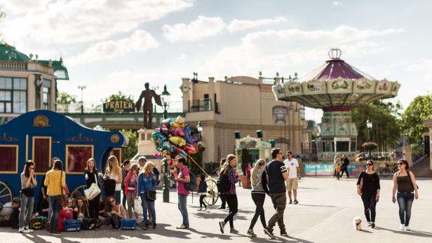Menschen flanieren im Wiener Prater vor einem Karussell und einer Statue.