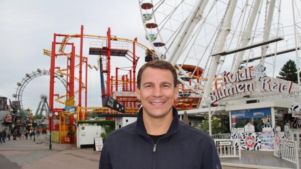 Ein Mann lächelt vor dem Wiener Prater mit Riesenrad und Achterbahn im Hintergrund.
