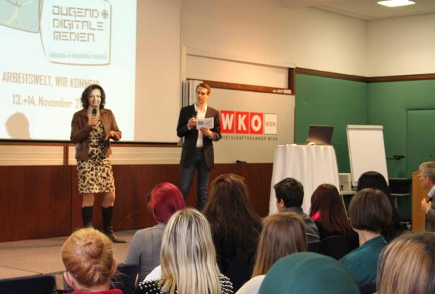 Eine Frau spricht auf einer Bühne vor Publikum, ein Mann steht daneben.