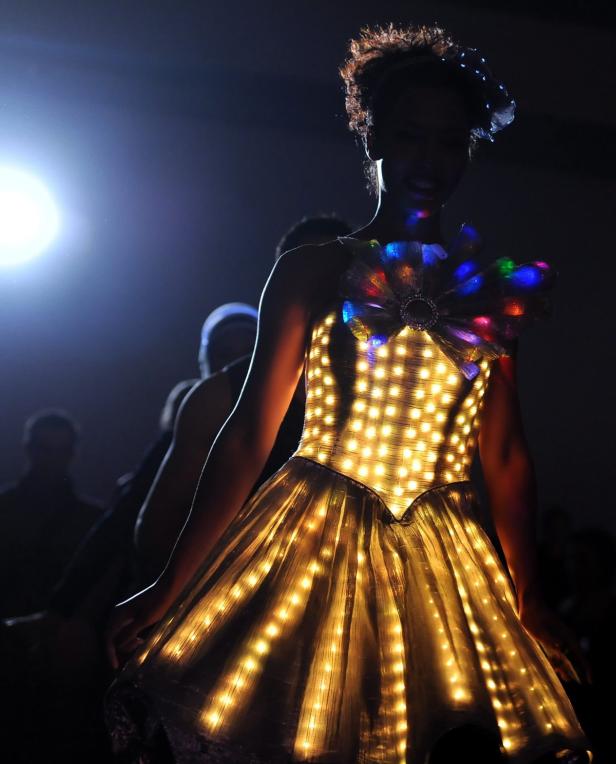 Eine Frau trägt ein leuchtendes Kleid mit bunten Lichtern auf einer dunklen Bühne.