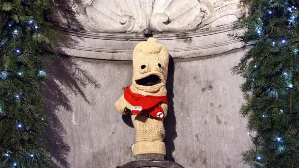 Manneken Pis in Brüssel mit einer gehäkelten Verkleidung und einer roten Schärpe, umgeben von Weihnachtsbäumen.