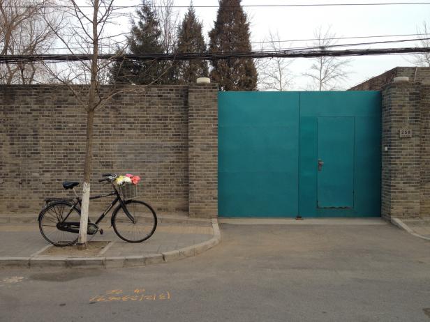 Ein Fahrrad mit Blumenkorb steht vor einer türkisfarbenen Tür in einer Backsteinmauer.