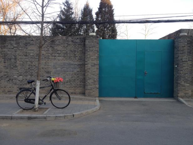 Ein Fahrrad mit einem Blumenkorb steht vor einer türkisfarbenen Tür und einer Backsteinmauer.