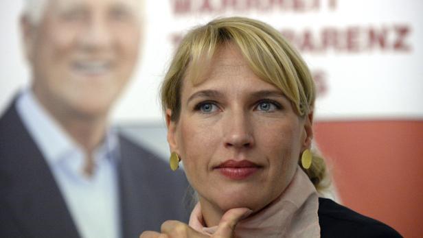 Eine Frau mit blonden Haaren und goldenen Ohrringen vor einem unscharfen Hintergrund.