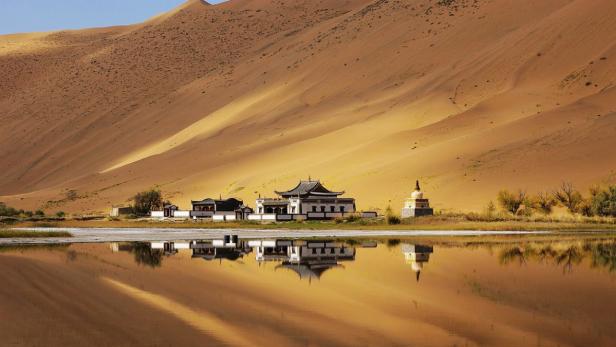 Ein buddhistischer Tempel und eine Stupa spiegeln sich in einem See vor einer Sanddüne.