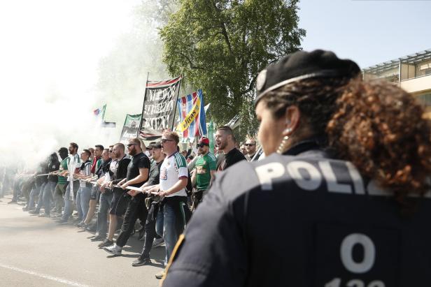Eine Polizistin steht vor einer Gruppe Fußballfans mit Fahnen und Rauch.
