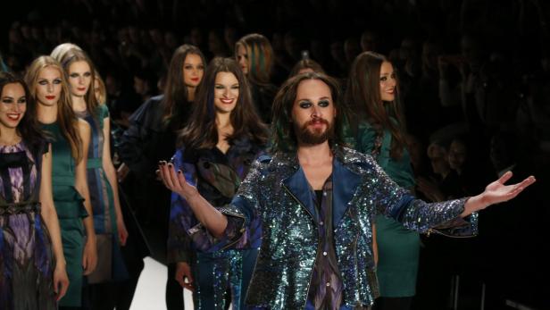 Ein Mann mit Bart und glitzernder Jacke steht auf einem Laufsteg mit Models.