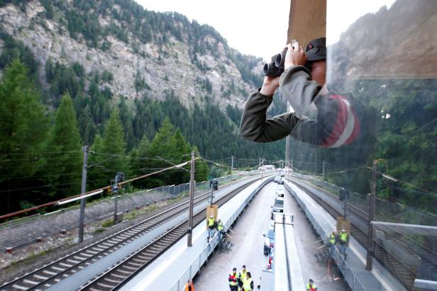 Eine Person blickt mit einem Fernglas aus dem Fenster auf eine Bahnstrecke in den Bergen.