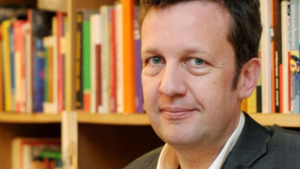 Ein Mann mit hellen Augen steht vor einem Bücherregal.