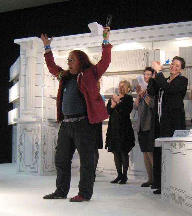 Ein Mann mit Mikrofon jubelt auf einer Bühne, während andere applaudieren.