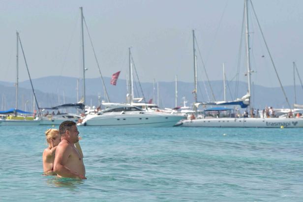 Ein Paar steht im türkisfarbenen Wasser, im Hintergrund Segelboote.