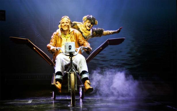 Ein Mann mit Fliegerbrille fährt ein seltsames Flugrad, während ein Mann im Katzenkostüm hinter ihm steht.