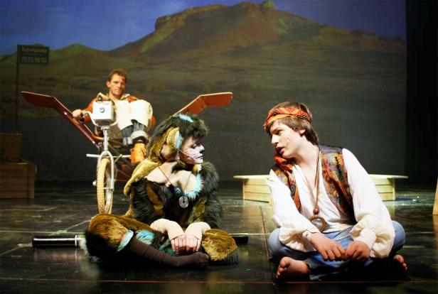 Zwei Schauspieler auf einer Bühne, einer im Kostüm, der andere mit Stirnband, im Hintergrund ein Mann auf einem Fahrrad mit Flügeln.